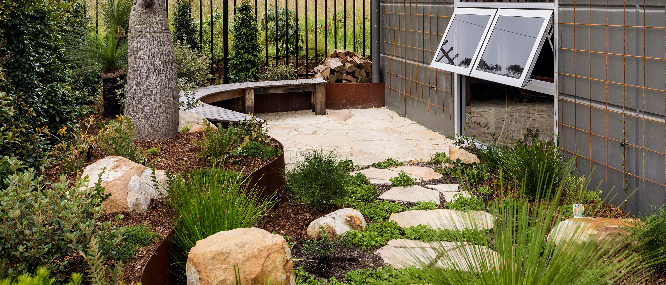 Meandering sandstone crazy pave stepper path through garden bed of dichondra and feature boulders to fire pit area with curved timber bench in courtyard